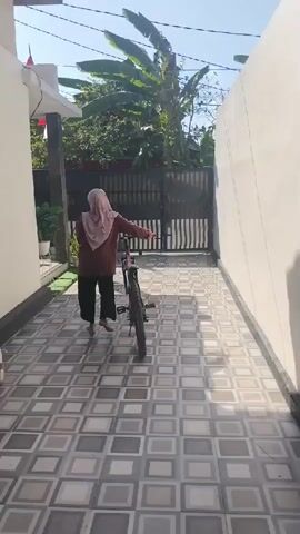 A person wearing a pink headscarf and a maroon shirt, along with black pants, is walking a bicycle towards a black metal gate. The person is positioned in the center of the frame, facing away from the camera and toward the gate. The bicycle is between the person and the gate. The ground is paved with square tiles in a checkerboard pattern. To the left of the frame, there is a white wall and a building with a red flag and some potted plants. To the right, there is a tall, smooth, light-colored wall. In the background, beyond the gate, are green trees and vegetation, and the sky is visible. The lighting suggests it is daytime, with sunlight casting shadows.