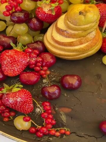 A close-up, overhead shot shows a dark, cracked surface adorned with an assortment of fresh fruits. A sliced pear, fanned out to reveal its segments, stands prominently at the top center. Surrounding the pear are bright red strawberries, bunches of green grapes, and deep red cherries. Sprigs of fresh mint are interspersed among the fruit. Scattered across the dark surface are individual grapes and cherries, adding to the decorative presentation. The overall scene suggests a dessert or a platter prepared for serving.