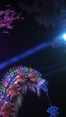This is a vertical video taken at night in what appears to be an outdoor, possibly rural, setting in Dukuh Klopo, Indonesia. The scene is dominated by a large sound system setup, adorned with festive decorations and illuminated by numerous flashing lights in vibrant purple, blue, and white. The sound system features stacked speakers and a prominent, decorative structure with a graphic that seems to depict a stylized figure or emblem. The overall atmosphere is that of a lively event, possibly a festival or celebration, with a crowd of people gathered around the sound system.

In the foreground, a person is seen walking away from the camera, wearing a plaid shirt. Other individuals are visible in the background, some of whom appear to be interacting with the sound system or enjoying the event. Trees with foliage frame the top and sides of the scene, suggesting the event is taking place outdoors, possibly near a road or pathway.

A notable element in the earlier part of the video (around 00:00-00:03) is a decorative structure resembling a dragon's head, also illuminated with colorful lights. This suggests a cultural or traditional aspect to the celebration, possibly related to local folklore or festivals. The presence of a red and white flag (around 00:01-00:03) might indicate a national celebration or a local event associated with Indonesia.

The visible text includes "RBX" and "RAGA KULI" on the central graphic of the sound system, and potentially "FEX" on one of the speakers. The lighting suggests a nighttime event, with a festive and energetic mood conveyed through the flashing lights and the gathering of people.