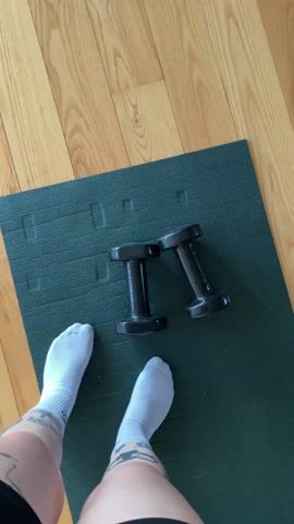Here's a detailed description of the video:

The video begins with a close-up shot of a green exercise mat on a wooden floor. Two black dumbbells rest on the mat. A person's feet, clad in white socks, are visible at the bottom left of the frame. 

The camera then pulls back, showing more of the wooden floor and a black bag near the dumbbells. The camera pans toward a large glass door with multiple panes.

The final shot focuses on the view through the glass door. It reveals a patio or balcony with potted plants, a railing, and a view of a green, hilly landscape with scattered houses. The overall scene suggests a peaceful, possibly rural or suburban, setting.