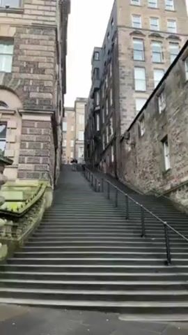 The video shows a long, stone staircase ascending between two tall buildings. The buildings are made of light-colored stone, with many windows. The staircase has metal railings on both sides. A person with an umbrella is walking up the staircase. The sky is overcast. The video is taken from a low angle, looking up at the staircase. The buildings appear old, with some moss growing on the stone. The street surface is a combination of stone and asphalt.