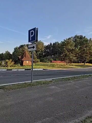 The video shows a road with a parking sign and an arrow pointing to the right. There are trees and a building in the background. The weather appears to be clear and sunny. The video is filmed from a stationary position, likely from the side of the road.