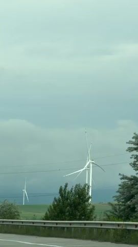 The video shows a view from a vehicle driving on a highway. Along the roadside are wind turbines in a field, and power poles. The sky is overcast with dark, cloudy weather.