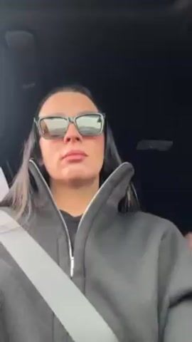 A woman with long dark hair, sunglasses, and a black zip-up sweatshirt is in the driver's seat of a car. She is wearing a seatbelt. Music is playing in the background. She smiles briefly. The interior of the car is visible, including the rearview mirror and the head of a small child in the back seat.