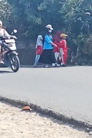 A man is riding a black scooter down a paved road. He is wearing a white shirt with red accents and grey shorts. He has grey hair and is not wearing a helmet. Behind him, on the side of the road, a group of people are walking. Several of them are wearing red shirts and headscarves. The environment appears to be outdoors with trees and foliage visible in the background. The road is bordered by a rough, gravelly shoulder. Another red scooter carrying two people passes by in the background as the first scooter continues down the road.