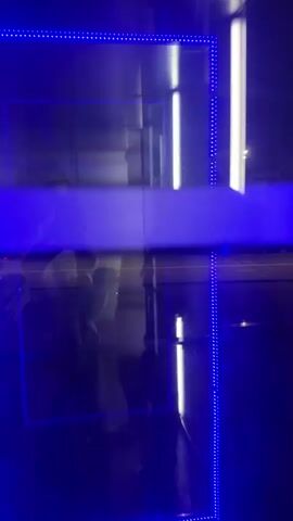 A person taking a photo of themselves in a reflective blue-lit booth. The person is partially visible in the lower left quadrant.  The lights inside the booth are reflected on the glass. Cars can be seen passing by on the other side of the glass, along with streetlights.