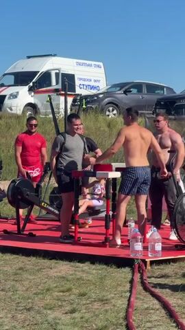 A group of men are gathered around an arm wrestling table set up outdoors on a red platform. One man in a red shirt stands to the left, facing the table. Two men are positioned at the table, facing each other with their hands clasped, preparing to arm wrestle. Another man stands behind them to the right, observing. A white van with "СПАСАТЕЛЬНАЯ СЛУЖБА" (Rescue Service) and "ГОРОДСКОЙ ОКРУГ СТУПИНО" (City District Stupino) written on it is visible in the background, along with other vehicles and trees. The ground is covered in grass, and the sky is clear and blue. There are two water rowing machines on the red platform.