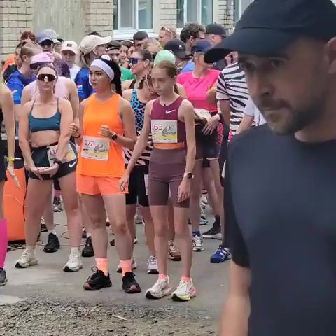 The video shows a group of people standing in what appears to be the start of a race. They are lined up facing forward, and many are wearing athletic clothing and bib numbers. The environment is outdoors, with a building visible in the background. Some individuals are looking down or to the side, possibly preparing for the race. There is a slight motion blur throughout the video, suggesting it was recorded while someone was walking or moving. The lighting indicates it is daytime.