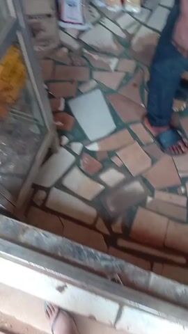The camera pans inside a small convenience store with tiled floors. A person in blue jeans and sandals is standing near the doorway. The store has shelves stocked with various items, including snacks, notebooks, and other goods. A man in a patterned shirt is standing near the counter. There is a glass display case filled with items on the counter.