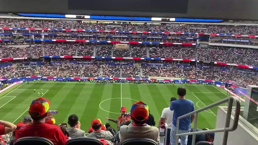 This video captures a high-angle view of a soccer stadium packed with cheering spectators. The scene is set in Inglewood, California, as indicated by the "LOS ANGELES" text visible on the stadium's upper levels. The stadium is brightly lit, suggesting it's daytime, and the weather appears clear, as there are no visible obstructions.

The primary focus is the large, illuminated screen above the field. This screen displays player information, featuring images of soccer players with their names and numbers, alongside the USA flag and the text "USA." The players shown are wearing soccer jerseys. One player is identified as "SEBASTIAN BERNHALTER" with the number 14. Another player, with the number 2, is also featured. The text "FIFA WORLD CUP 2026" is also visible on the stadium's seating.

The soccer field is a vibrant green, marked with white lines for the game. A few players are visible on the field, appearing to be warming up or preparing for a match. In the foreground, the backs of spectators are visible, many wearing red shirts and some with colorful hats, their focus directed towards the field and the giant screen. The overall atmosphere is one of excitement and anticipation, typical of a major sporting event. The crowd appears enthusiastic, with their attention captivated by the pre-game proceedings.