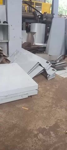 The scene shows an industrial workshop filled with various fabricated metal components. Dominating the foreground is a stack of rectangular panels painted white. Some have identifying marks scrawled on their surfaces. Scattered around are other pieces of metal, mostly flat and angular, suggesting they are in-progress parts or cutoffs. A few components resemble incomplete box shapes. The background contains industrial machinery, including a large yellow piece of equipment and cylindrical metal structures. The floor is concrete and covered with debris, adding to the messy, utilitarian feel of the workshop. There are no people visible in the video.