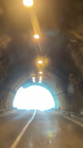 The video shows a point-of-view shot of a car driving through a tunnel and then emerging into a mountainous landscape. Inside the tunnel, the walls are rough and made of stone or concrete. Lights are mounted on the ceiling at intervals. As the car approaches the end of the tunnel, the bright daylight becomes visible. Once outside the tunnel, the view opens up to a scenic vista of green mountains, some of which are capped with snow. The sky is blue with scattered white clouds. The road is paved and has white lane markings.