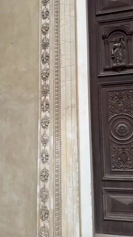 A close-up, medium shot shows a section of an ornate building facade, focusing on a large, dark wooden double door with intricate carvings. To the left of the door is a pale, textured wall. Adjacent to the door, there is a column-like architectural element with decorative plasterwork, featuring a vertical band of repetitive motifs resembling clusters of grapes and leaves. This decorative band is bordered by two narrower bands, one with a repeating geometric pattern and another with a more textured appearance. The area around the door frame and the plasterwork shows signs of age and wear, with some cracks and discoloration visible. Above the door, a decorative cornice with detailed carvings is present. The video pans slightly upward and to the right, revealing more of the upper architectural details and the transition to a yellow-toned upper section of the building. The lighting is even, suggesting an outdoor setting during the daytime. No people are visible in the frame.