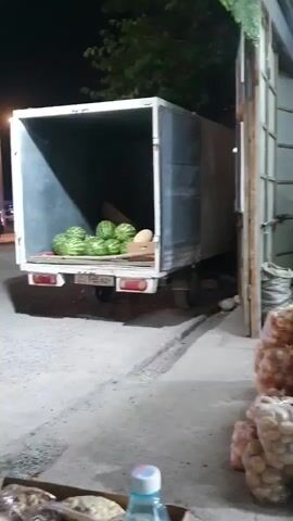 The video opens on a nighttime scene at what appears to be a fruit and vegetable market. In the foreground, several large bags of produce, possibly potatoes, are stacked on the ground. Behind these, there are shelves overflowing with various fruits and vegetables in crates and bins. To the left, the open back of a truck is visible, loaded with what looks like watermelons. The market stalls are lit by artificial lights, creating a somewhat dimly lit atmosphere. The camera pans slowly across the scene, showing different sections of the market. The environment appears to be an open-air or semi-enclosed space, with corrugated metal walls and a roof visible. The overall impression is one of a busy market at the end of the day or before opening.