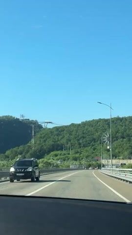 This is a POV video captured from a vehicle driving on a highway. The highway has multiple lanes and is bordered by white guardrails with red reflectors. On the left side of the highway, there is a lush, green mountainside covered in trees. A tall tower with a white structure at its peak, possibly a ski lift or zipline, is visible on the mountainside. Power lines and poles are also present. On the right side of the highway, more green mountains are visible, and a large decorative circular object resembling a metal windmill or starburst is attached to a lamppost. Further down the road, the mountainside shows some signs of construction or quarrying. The sky is a clear, bright blue, indicating a sunny day. The overall mood is peaceful and scenic, suggesting a pleasant journey through a natural landscape. The location is identified as Kazachiy Brod, Russia. The time of day appears to be daytime, likely late morning or afternoon, given the bright sunlight. There is no visible text in the video.