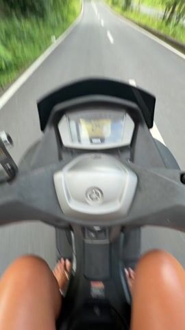 A woman is riding a Yamaha scooter on a paved road lined with green vegetation. The video is shot from a first-person perspective, showing the scooter's dashboard indicating a speed of 24. The woman is wearing a yellow and white striped shirt, dark sunglasses, a necklace, and sandals. She is looking at the camera and smiling. The road has a white dividing line.