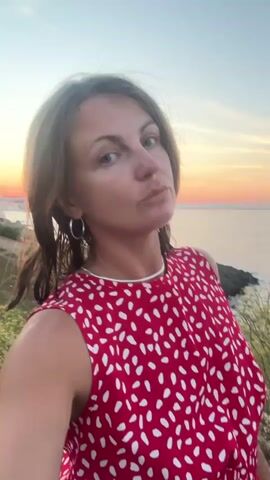 The video shows a woman in a red and white polka dot dress standing outdoors near a body of water, possibly the sea. The sky behind her is a warm gradient of orange and blue, indicating it is either sunrise or sunset. The woman is looking at the camera, with her head tilted. She has brown hair and is wearing hoop earrings. The background includes rocky terrain and vegetation. The woman makes a kissing face toward the camera at one point.