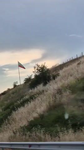 The media file shows a video taken from a moving vehicle, likely a car, looking towards a grassy hillside. The dominant subject is a flag flying from a flagpole on the crest of the hill. The flag has three horizontal stripes: white on top, green in the middle, and red on the bottom. Behind the flag, the sky is cloudy with patches of lighter sky suggesting a sunset or sunrise. The hillside is covered in dry grass and some patches of green vegetation, with a few scattered trees and bushes. A fence is visible running along the top of the hill behind the flag. The overall scene is natural and rural. The time of day appears to be late afternoon or early evening due to the lighting and cloud cover. The weather seems calm, with the flag gently waving. There is no visible text in the media file. The location context indicates the scene is in Peshterna, Bulgaria, and the flag is likely the Bulgarian flag. The mood conveyed is one of patriotism and possibly a sense of journey or passage.