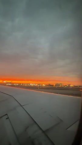 The video shows a view from an airplane window during takeoff or landing. The sun is setting, casting a vibrant orange glow across the sky, which is partially covered by gray clouds. In the distance, the lights of an airport or city are visible. The wing of the airplane is prominent in the foreground, stretching across the frame and leading the eye towards the horizon. The wing appears to be clean and well-maintained, with navigation lights visible along its edge. The overall scene conveys a sense of travel and the beauty of a sunset from high above the ground. There are no people visible in the video.