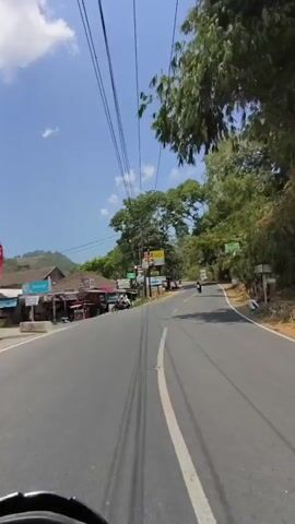 A first-person perspective video shows a person riding a motorcycle on a paved road. The road is surrounded by lush green trees and vegetation. The sky is blue with a few scattered clouds. The motorcycle passes by small buildings and signs along the side of the road. In the distance, another motorcycle is visible. The rider's hands are visible on the handlebars. The video appears to be filmed during the daytime.
