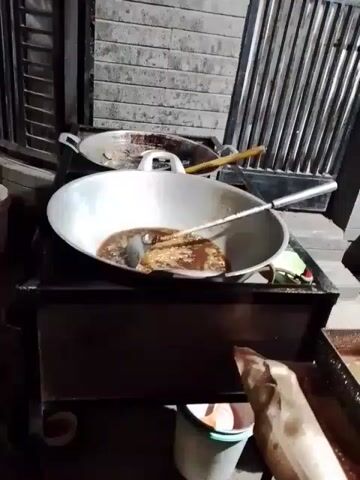 A video captures a large metal wok sitting on a dark-colored cooking station. Inside the wok is a dark liquid bubbling with something frying. A long, metal ladle rests in the wok. Behind the wok is another pan, also on the cooking station, along with some wooden cooking utensils. A white bucket is underneath the cooking station. The background includes a wall with a brick-like pattern and a barred window or fence. A person's arm and torso are briefly visible in the lower right corner of the frame, clad in a tan garment. The scene appears to be outdoors at night.