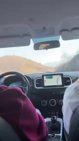 The video captures the perspective from inside a car driving along a winding mountain road. Two people are visible in the front seats: the driver wearing a maroon hoodie and the passenger in a white sweatshirt. A GPS screen displays the route ahead. The scenery outside is characterized by dry, rocky hills under a clear sky. The car's dashboard is visible, showing controls for temperature and other functions. The overall mood is that of a road trip through a scenic, natural landscape.