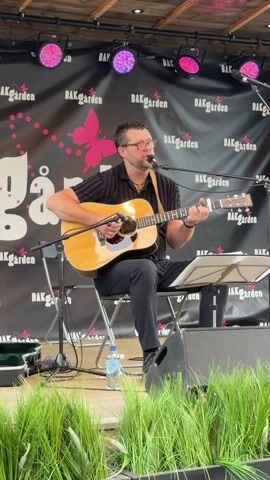 A man with glasses and a striped shirt plays an acoustic guitar and sings on a stage. He is sitting on a metal chair, and there is a microphone in front of him. A banner with the text "BAKGården" and a butterfly image is behind him. The stage appears to be outdoors, with some green plants in the foreground. There are lights above the stage.
