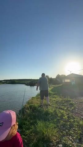 The video shows a little girl in a pink t-shirt and baseball cap standing by a body of water.  Behind her, a man in a green shirt and shorts is fishing. The sun is shining brightly in a clear blue sky. The environment appears to be rural, with green grass and a few structures in the background. The man casts his fishing line into the water. The little girl seems to be watching him and talking.