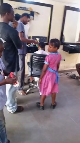 The video shows the interior of a barber shop with several people inside. A young girl in a pink and white checkered dress with a blue ribbon tied at the back stands in the center, looking slightly over her shoulder. A young boy in a white t-shirt and jeans walks past her. To the left, a barber wearing a black shirt and light-colored pants is working on a child's hair, who is seated in a barber chair. In the background, another man is getting his hair cut. A man in a blue shirt is sitting in a chair and looking at his phone. There is a second young boy running around in the background. The shop has mirrors on the walls and tiled floors.