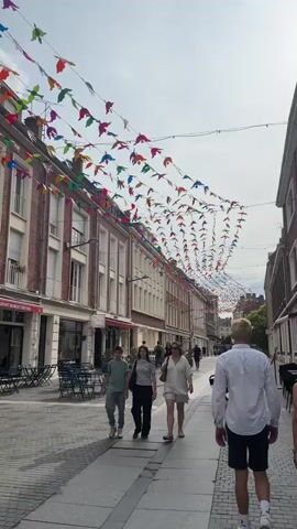 The video shows a street scene with buildings lining both sides. Colorful bird-shaped decorations are strung across the street overhead, creating a festive atmosphere. People are walking along the paved street. In the foreground, a man with blonde hair wearing a white shirt and dark shorts walks away from the camera. Behind him, a woman with blonde hair wearing a white outfit walks with her back to the camera, carrying a dark bag. Further down the street, two people, a younger male in a green shirt and a female in a striped shirt, walk towards the camera. Outdoor seating with tables and chairs is visible along the sides of the street, some of which are empty. The buildings have multiple stories with windows and shutters. The sky is overcast.