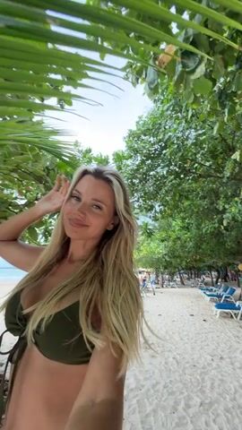 A woman with blonde hair and a green bikini smiles and poses for a selfie on a sandy beach. She is partially obscured by a large green palm frond in the foreground. Behind her, palm trees and other foliage line the beach, and lounge chairs are visible in the distance. The woman adjusts her hair and looks at the camera, smiling and touching the palm frond.