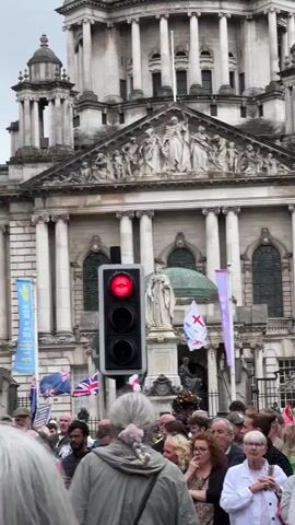 This is a medium shot video of a crowd of people standing in front of a large, ornate building. A traffic light in the foreground is displaying a red light. Several flags are visible, including Union Jack flags and banners with religious symbols. The people in the crowd appear to be diverse in age and ethnicity, and they are looking towards the building. The building itself is classical in style, with columns and intricate carvings. The sky is overcast.