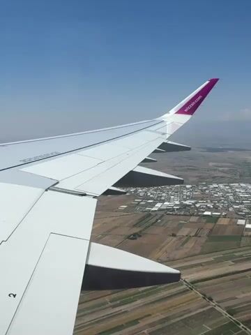 The video shows a close-up view of an airplane wing from inside the cabin. The wing is white with a purple tip. The sky is blue and clear, and the ground below is a patchwork of fields and buildings, suggesting the plane is in flight over a rural or semi-urban area. The footage is slightly blurred, especially in the distance, possibly due to motion or a slightly out-of-focus camera. The scene appears to be captured during daylight hours.