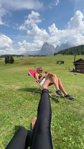 A woman is sitting in a red lawn chair outdoors, holding a phone. In the foreground, another person's legs are visible, wearing black leggings. The background shows a grassy field with mountains and a blue sky with white clouds.