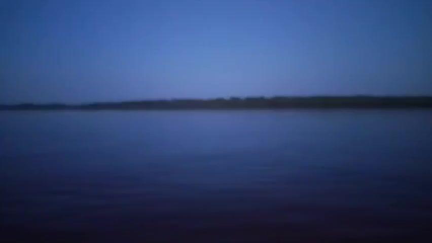 The video begins with a blurry shot of a dark body of water under a dark blue sky. The image then shifts to a slightly clearer view of the water and a sandy shore. As the video progresses, the scene becomes sharper, revealing a wide river or large body of water. On the left side of the frame, a paved walkway lines the shore, illuminated by streetlights. Several people are walking along the pathway. Buildings and structures, including what appears to be a tall building, are visible in the distance along the shoreline. The sky remains a deep blue, indicating either early morning or evening twilight. The overall atmosphere is calm and peaceful.