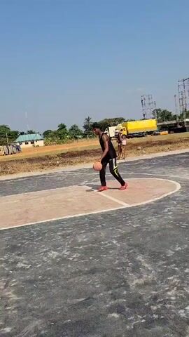 On a basketball court, a dark-skinned person wearing a black tank top, black athletic pants with white and yellow accents on the side, and red sneakers is observed taking a shot. The basketball is orange and brown. The court is grey with a beige free-throw lane. A basketball hoop is present. The shot is missed, bouncing off the rim. The background includes a light blue sky, trees, a building, and a yellow truck in the distance.