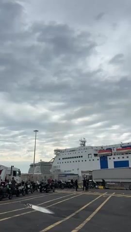 The video shows a parking area at a port, with a large white and blue ferry docked in the background. The ferry, named "GNV Auriga," has the words "Lillo Palermo & Figli" printed on the side of a red trailer parked nearby. The sky is overcast with gray clouds. In the parking area, there are rows of parked motorcycles and cars. People are milling around the vehicles, some appear to be loading or unloading. A large cruise ship is partially visible in the background on the left side of the frame. The scene suggests preparation for boarding the ferry or disembarking.