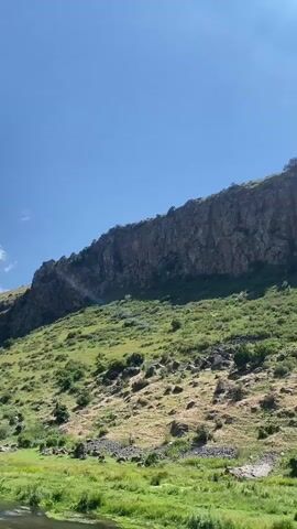 The video shows a large, rugged rock formation against a clear blue sky. The rock face is steep and textured, with some vegetation growing on its lower slopes. Below the rock formation, there is a green, grassy area with scattered rocks and bushes. In the foreground, there is a body of water, likely a river or stream, with lush green vegetation along its banks. The sun is shining brightly, casting shadows and highlighting the landscape. The video pans slightly to the right, revealing more of the rock formation and the surrounding natural environment.
