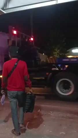 A person wearing a red long-sleeved shirt, blue jeans, and a black cap walks away from the camera towards a large flatbed truck at night. The person is carrying a black bag in their right hand and a purple object in their left hand. The truck has multiple large wheels and is partially illuminated by streetlights and the truck's own lights. The truck appears to be in motion, as indicated by the blur on its wheels. In the background, there are trees, a building with lights, and other vehicles, including motorcycles and a white SUV, that are also in motion. A red traffic signal is visible in the upper left portion of the frame. The ground is paved and appears wet in some areas.