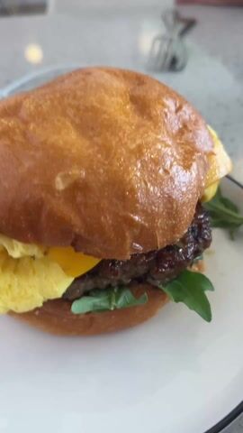 Here's a concise description of the video: 

A close-up shot showcases a breakfast sandwich on a white plate with a thin black rim. The sandwich has a shiny brioche bun, a sausage patty, arugula, yellow cheese, and scrambled eggs. The plate sits on a light-colored speckled table in what appears to be a restaurant or cafe. Other people are seated at the table, partially visible in the background.