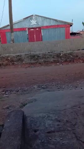 The video shows a building with a silver metal roof, blue-gray siding, and red trim around the top and the door, which has a white dove painted on its front. A person wearing a dark shirt with light-colored leaves walks along the top of a cinderblock wall that runs in front of the building. The wall separates the building from a red dirt road in the foreground.  Another person walks behind them, holding a piece of white paper. A dark blue car drives by on the dirt road to the left. There is a utility pole to the left of the building. A large, gray rock is in the foreground to the left.