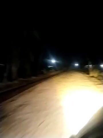 The video shows a first-person perspective of someone driving down a dirt road at night. The road is illuminated by the vehicle's headlights, and there are occasional streetlights in the distance. The surrounding environment is dark, with some trees and structures visible on either side of the road. The camera is shaky, indicating movement, and there are no other people or vehicles visible in the frame.