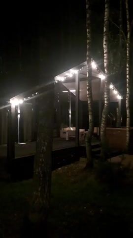 The video shows a nighttime view of two structures in a wooded area. The first structure, on the left, appears to be a modern building with a transparent roof and interior lighting. String lights are visible along the edge of the roof. The second structure, on the right, is a smaller, darker building with visible windows showing interior light. Trees are present in the foreground and background, obscuring some of the view. The overall scene is dark, with the artificial lights of the buildings being the primary illumination.
