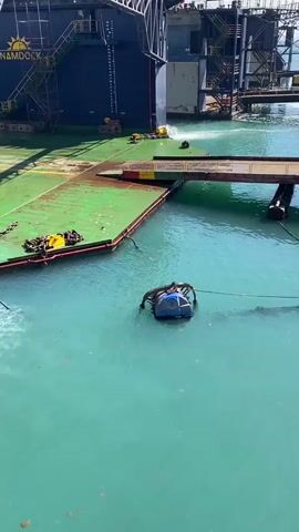 The video shows a large, dark blue ship labeled "NAMDOCK 3" docked in a harbor. The dock has a green platform. A small, blue, robotic device floats in the turquoise water near the dock. Water sprays from a pipe into the water. Overhead, cranes stand tall against a clear, blue sky. Some people are visible on the ship.