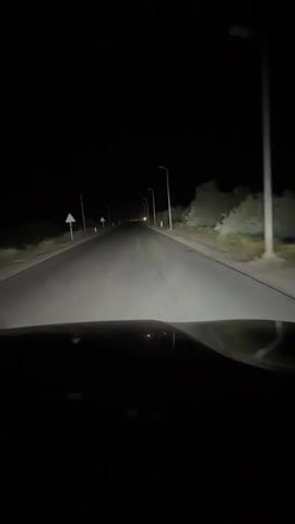 This is a first-person view from inside a vehicle driving on a dark, paved road at night. The headlights of the vehicle illuminate the road ahead, revealing a straight path with white lane markings. There are streetlights visible along the side of the road, and sparse vegetation and trees can be seen in the distance. The sky is completely dark, indicating nighttime. There are no other vehicles or pedestrians visible in the immediate vicinity. The dashboard of the vehicle is partially visible in the foreground. The scene suggests a rural or suburban setting due to the lack of dense development and the presence of open space.
