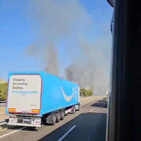 A bright blue Amazon Prime semi-trailer truck is stopped on a highway, with thick black smoke billowing from behind it and further down the road. The truck has the Amazon Prime logo on its side and text on the back that reads "Shipping. Streaming. Savings. It's on amazon prime." Another semi-trailer truck is visible to the right, also stopped. The sky is clear and blue, and the highway appears to be in a rural or suburban area with trees and vegetation visible.