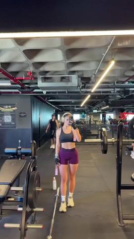 A young woman with brown hair, wearing a black sports bra, purple shorts, white socks, and athletic shoes, stands in a gym. She is wearing white headphones and holding a smartphone in her right hand, taking a mirror selfie. In her left hand, she holds a pink resistance band. A water bottle is on the floor next to her. In the background, a man wearing a black tank top, black shorts, and a black baseball cap is walking away from the camera. The gym has black rubber flooring, metal lockers, weight racks with barbells and weights, and fluorescent lighting on the ceiling.