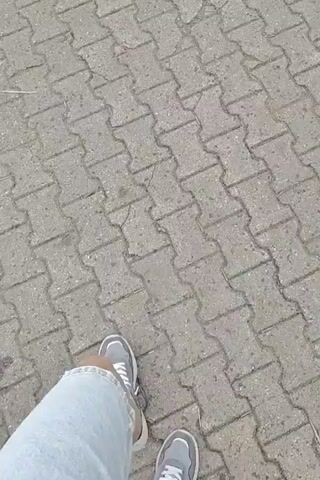 This is a first-person perspective video of someone walking on a paved walkway. The walkway is made of interlocking concrete pavers arranged in a herringbone pattern. The person is wearing grey and white sneakers and light blue denim shorts. The camera is pointed downwards, focusing on the ground and the person's legs and feet as they walk. The lighting suggests it is daytime, likely outdoors, with an overcast sky given the soft, diffused light and lack of strong shadows. The scene is simple and mundane, depicting the act of walking. There is no visible text in the video. The context provided indicates the location is Palmiry, Poland, which could suggest a park, a street, or a similar outdoor public space within that area. No specific emotions are conveyed, other than the general sense of movement and progression.