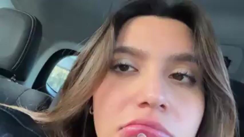 A close-up shot of a young woman sitting in what appears to be a car. She has long brown hair and is wearing a black leather jacket. She is looking up and to the side. Her lower lip appears swollen, and she is touching it with her finger, seemingly examining it. The background is slightly blurred, but the car's interior, including a headrest, is visible.
