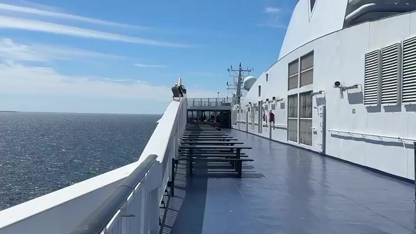 The video shows a view from the deck of a large white ferry sailing on a vast expanse of dark blue water under a bright blue sky with scattered white clouds. The ferry's railing and deck are visible in the foreground, with rows of outdoor picnic tables and benches. The camera pans to reveal the open sea, with a distant, hazy shoreline visible on the horizon.

A second ferry, larger and with a distinctive dark and white hull and a green stripe, sails across the screen from left to right. The text "ECKERÖLINE" is clearly visible on its side, identifying it as part of the Eckerö Line, a ferry company operating in the Baltic Sea, primarily between Finland and Estonia. The distant shoreline appears to be the coast of Estonia.

The weather is sunny and fair, suggesting a pleasant day for travel. The bright sunlight reflects off the water, creating a shimmering effect. The overall mood is peaceful and expansive, evoking a sense of journey and open space. The time of day appears to be daytime, likely late morning or early afternoon, given the strong sunlight and clear sky. There are no people visible in the foreground or on the deck of the ferry the video is taken from, suggesting a quiet moment on board. The activity is primarily the ferry's journey across the sea.
